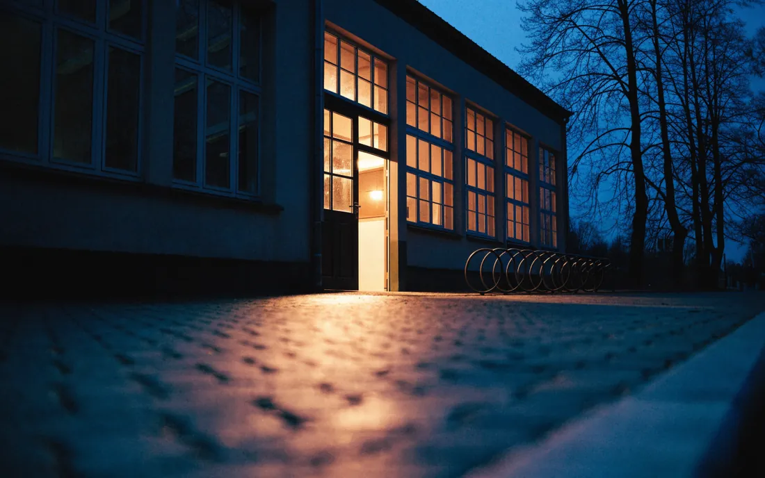 Szkoła o zmierzchu z zewnątrz: świecą wyłącznie okna sali gimnastycznej, boczne wejście otwarte, reszta budynku ciemna.
