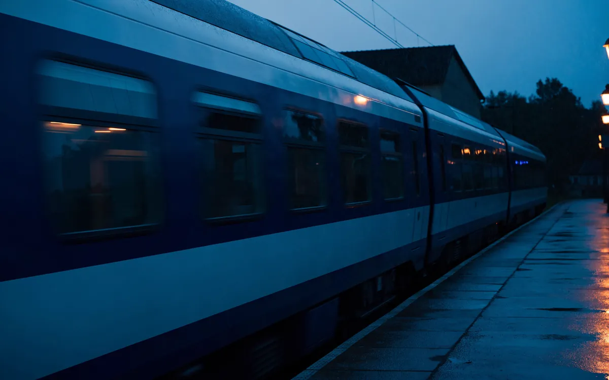 Pociąg podmiejski przy peronie o zmierzchu, mokra nawierzchnia i zapalone latarnie.